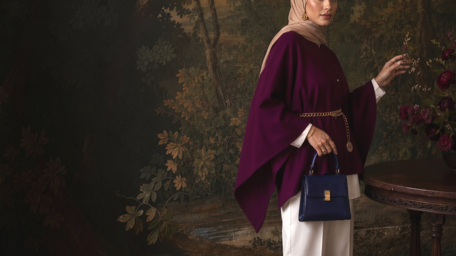 A woman cropped just below the eyes, in a plum wool cape over a cream shirt and trousers, a gold chain belt at the waist and a navy top-handle bag in her hand, against a dark painted woodland backdrop