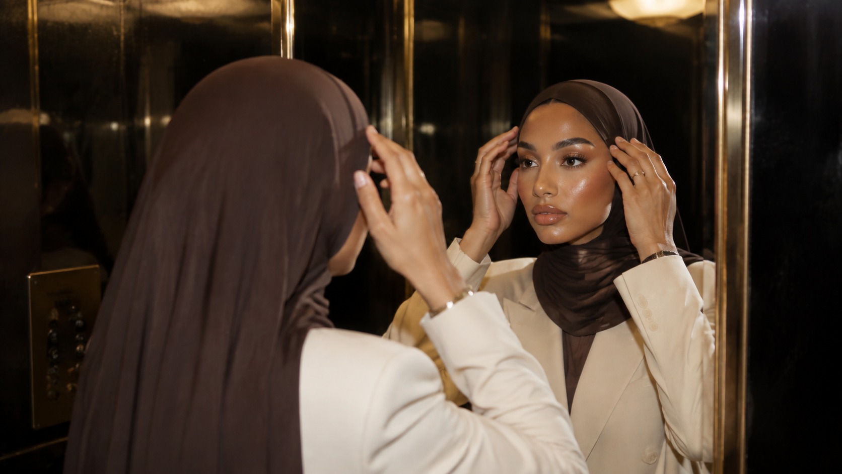 A woman adjusting a brown jersey hijab in a gold mirrored lift, wearing a cream blazer