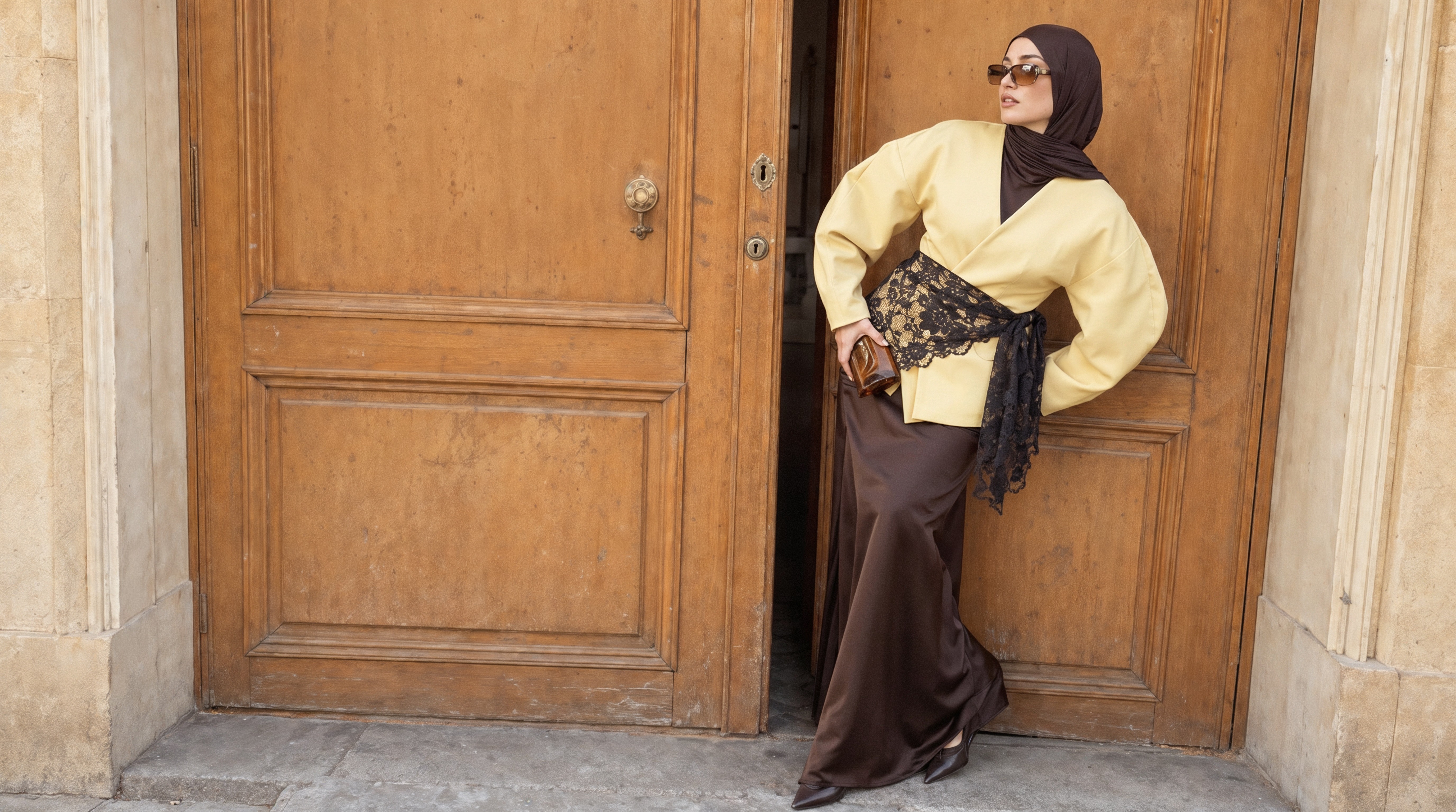 A woman in a brown hijab and sunglasses leaning in a doorway, wearing a black lace sash tied over a butter-yellow jacket and a brown satin maxi skirt