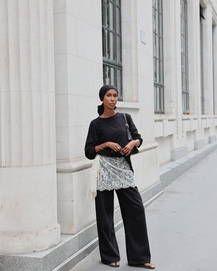 A woman in a plain black top and black wide-leg trousers with a white lace overskirt at the hip