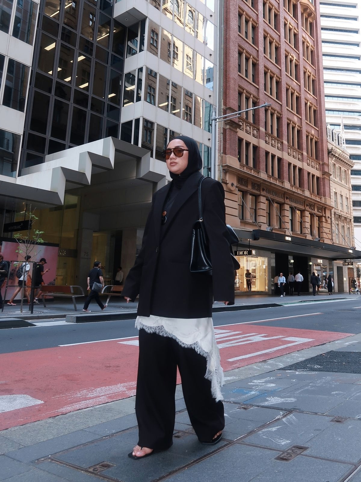 A woman in an oversized black coat and black trousers with a white lace skirt showing beneath