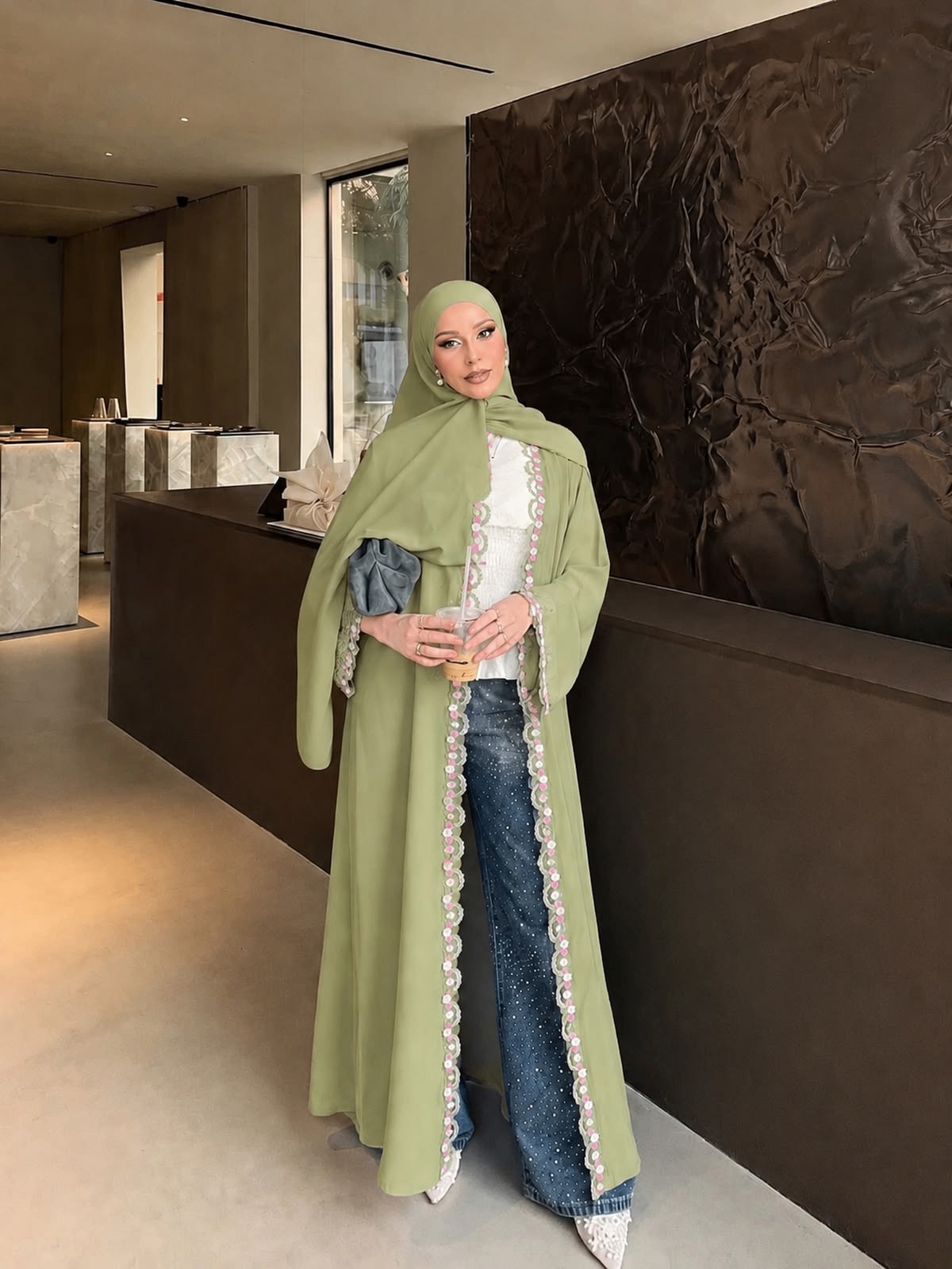 A woman in a sage green open abaya with floral lace trim down the front, worn over jeans